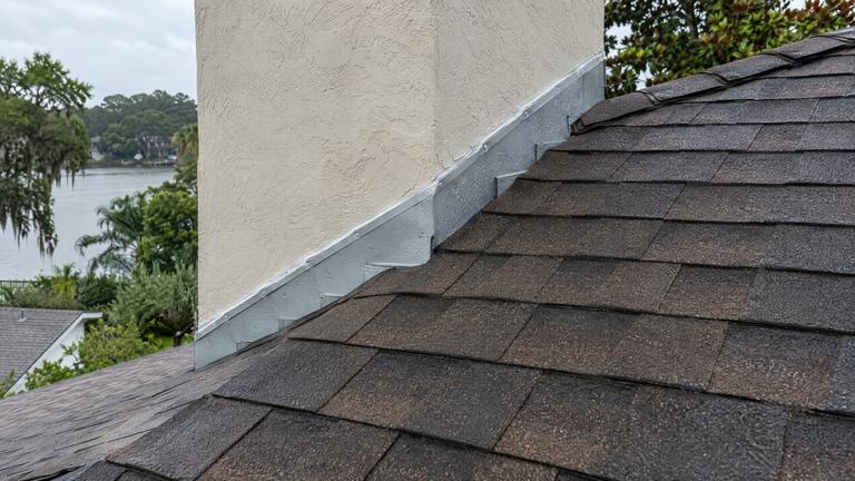 New metal flashing where a stucco chimney meets a shingle roof, with raindrops on the metal.
