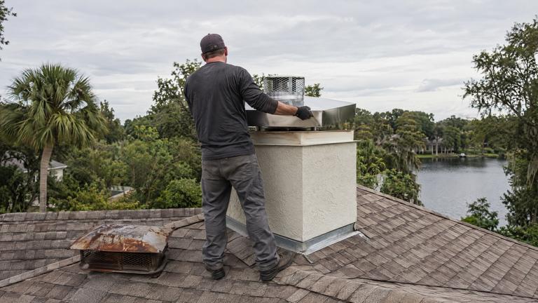 A worker lifts a new stainless chase cover onto a stucco chimney chase, the old rusted cover lying on the shingles.