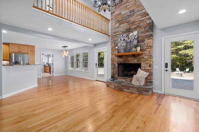 A floor-to-ceiling stacked stone fireplace with a wood mantel and raised stone hearth in an open room with hardwood floors and a kitchen behind.