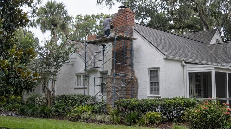 A tall brick chimney on a white stucco cottage being rebuilt from a steel frame scaffold.